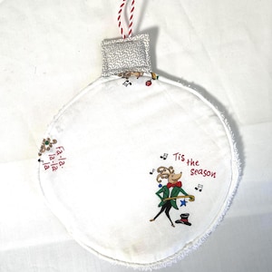 Peut inclure: Ornement de Noël en tissu blanc avec une boucle de suspension rayée rouge et blanche. L'ornement représente une souris de dessin animé en veste verte et nœud papillon rouge, avec le texte "Tis the season".