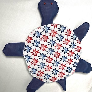 May include: A handmade turtle plushie with a navy blue body and limbs. The shell is patterned with red, white, and blue stars. The turtle has red bead eyes, and is a patriotic themed item.