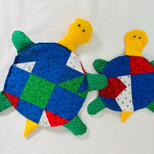 May include: Two handmade turtle-shaped plush toys. The turtles have blue, green, red, and white patchwork shell designs. They feature yellow heads and tails, and green flipper-like appendages. The toys are placed on a white surface.