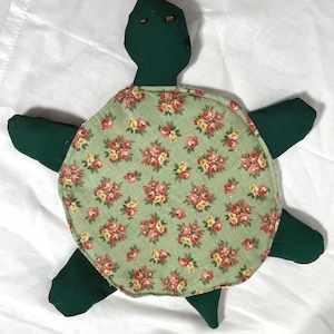 Puede incluir: Tortuga de tela verde hecha a mano con un caparazón estampado floral. El caparazón presenta un fondo verde claro con rosas rojas y amarillas. La tortuga tiene cuatro extremidades en forma de aleta y una cabeza con pequeños ojos de cuentas.