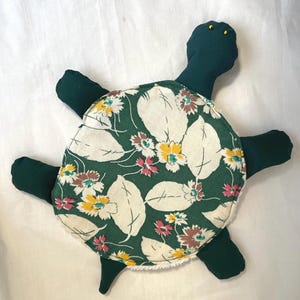 Puede incluir: Un peluche hecho a mano con forma de tortuga. El caparazón de la tortuga es de tela verde con un estampado floral de hojas blancas y flores rosas, amarillas y marrones. La cabeza, las patas y la cola son de color verde oscuro liso. La tortuga tiene ojos de cuentas amarillas.