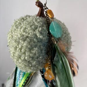 May include: A handmade, whimsical ornament featuring a light green, textured pom-pom shape. It is adorned with a brown stem, a chain with orange and teal beads, and colorful ribbons and feathers. The ornament is suspended by a green ribbon.