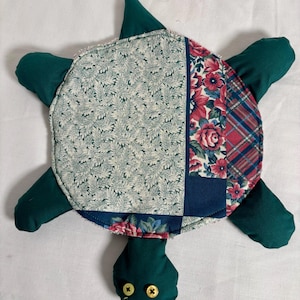 May include: A handmade turtle-shaped pillow with a patchwork shell. The shell features floral and plaid patterns in red, blue, and white, with a green leafy pattern. The turtle has green flippers, head, and a tail, with button eyes.
