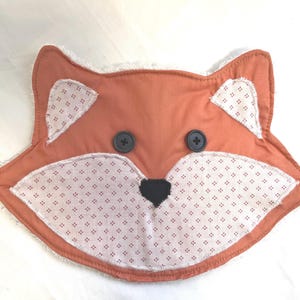 May include: A handmade, orange fox-shaped pillow with a white, patterned face and ears. The fox has two black button eyes and a black heart-shaped nose. The pillow's edges are trimmed with white, fluffy fabric.