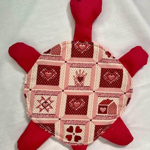 Könnte beinhalten: Ein handgefertigtes rotes Schildkröten-Plüschtier mit einem gemusterten Panzer. Der Panzer hat ein Patchwork-Design mit Herzen, Sternen und Handmotiven in Rot- und Rosatönen. Die Schildkröte hat einen langen Hals, vier Flossen und kleine schwarze Augen.