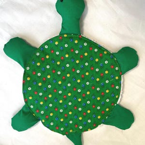 Könnte beinhalten: Ein handgefertigtes, grünes, schildkrötenförmiges Plüschtier. Die Schildkröte hat einen grünen Kopf, vier grüne flossenartige Gliedmaßen und einen grünen Schwanz. Der Panzer der Schildkröte ist ein Kreis aus grünem Stoff mit einem Blumenmuster aus roten, gelben, blauen und weißen Blumen.