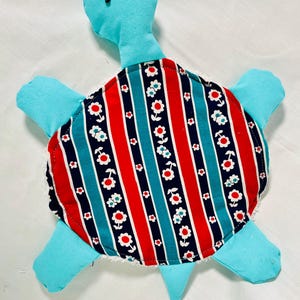 May include: A handmade stuffed turtle toy. The turtle has a light blue head, flippers, and tail. The shell is patterned with red and blue vertical stripes, interspersed with white and red floral designs. The turtle's shell is approximately 8 inches in diameter.