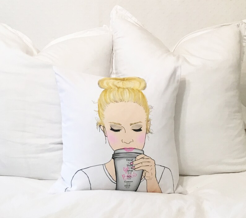 May include: A white pillow with a drawing of a woman with blonde hair in a bun, wearing a white shirt and drinking from a coffee cup. The coffee cup has the words "Decaf, Shots, Syrup, Milk, Cream, Custom Drink" written on it.