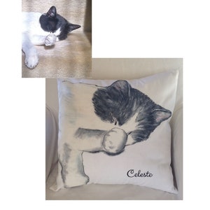 May include: A white and black cat with gray eyes is painted on a white pillow. The cat is sleeping with its paw covering its face. The pillow has the name "Celeste" embroidered on it in black cursive.