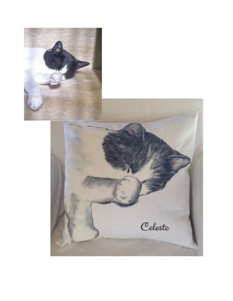 May include: A white and black cat with gray eyes is painted on a white pillow. The cat is sleeping with its paw covering its face. The pillow has the name "Celeste" embroidered on it in black cursive.