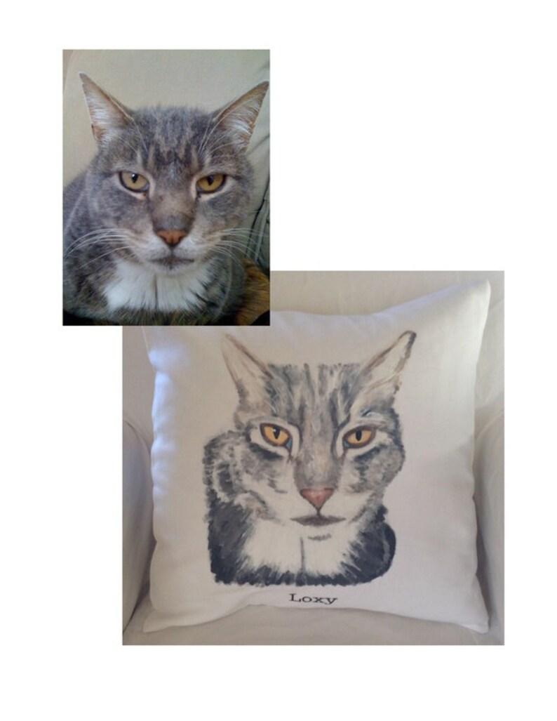 May include: A white pillow with a hand-painted portrait of a gray tabby cat with yellow eyes. The cat's name, "Loxy", is written in black lettering below the portrait.