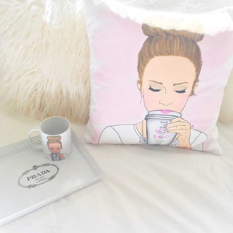 May include: A white pillow with a pink background featuring an illustration of a woman with brown hair in a bun, wearing a white shirt and a necklace, drinking from a white coffee cup with pink writing that says "Get Your Boss On".