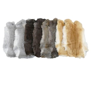May include: A collection of rabbit fur pelts in various natural colors, including shades of gray, brown, and tan. The pelts are arranged side by side, showcasing the soft texture and variations in the fur.