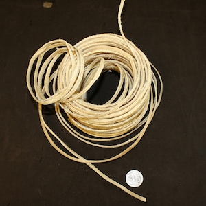 May include: A coil of off-white leather cord, approximately 1/4 inch wide, is wrapped around itself on a black surface. A silver coin is placed next to the cord.