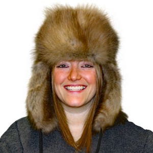 May include: A brown faux fur trapper hat with ear flaps. The hat is worn by a person with long brown hair and a smiling face.