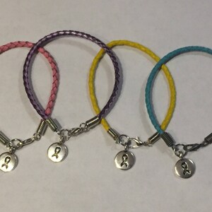 Pode incluir: Três pulseiras de couro trançado com pingentes de prata em forma de fita. As pulseiras são rosa, roxo e turquesa.