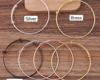 May include: Six different colored metal bangle bracelets on a wooden surface. The bracelets are silver, brass, bronze, platinum, rose gold, and gold.