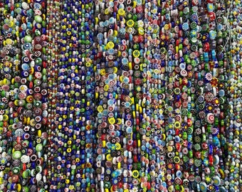 Puede incluir: Primer plano de una pared de cuentas de vidrio de colores, cada cuenta tiene un patrón y un color diferente. Las cuentas están enhebradas en largos hilos, creando una pantalla vibrante y texturizada.