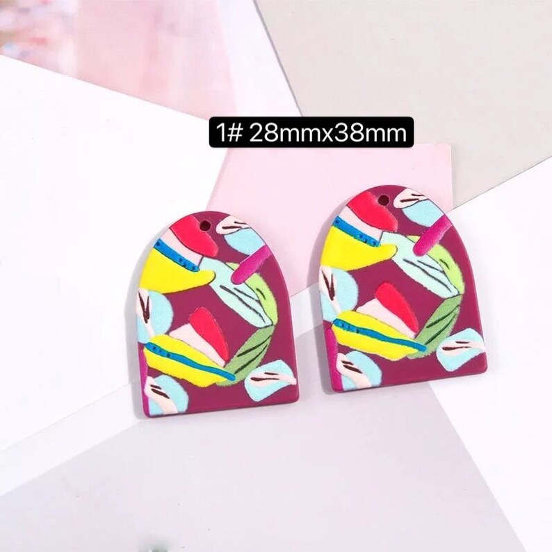 May include: Two colorful arch-shaped polymer clay earrings with a red base and abstract patterns in yellow, blue, green, and pink. The earrings are 28mm x 38mm.