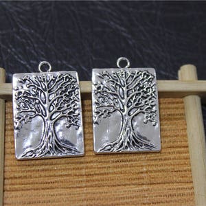 Puede incluir: Dos dijes de metal plateados en forma de rectángulos con un diseño de árbol. El árbol está detallado con ramas y raíces.