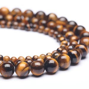 May include: Strands of polished tiger's eye gemstone beads in varying sizes. The beads are a mix of brown and gold, with a chatoyant effect. The beads are strung together, creating a curved shape against a white background.