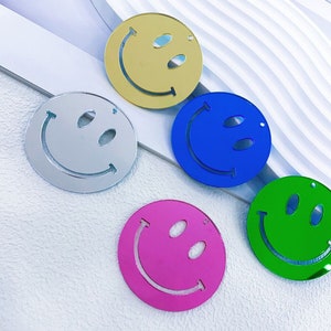 May include: Five different colored smiley face acrylic ornaments. The ornaments are gold, silver, blue, green, and pink. Each ornament has a simple smiley face design.