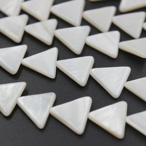 Puede incluir: Primer plano de cuentas triangulares blancas con brillo nacarado. Las cuentas están dispuestas en filas, cada una con esquinas redondeadas, creando un patrón sobre un fondo oscuro. Estas cuentas son adecuadas para la fabricación de joyas.