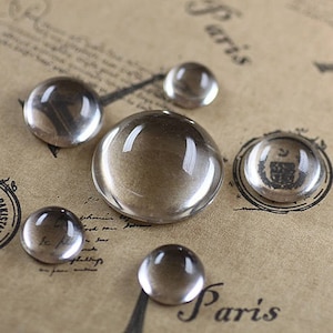 May include: Five clear glass cabochons of varying sizes, arranged on a brown paper surface with the word "Paris" written in black ink.