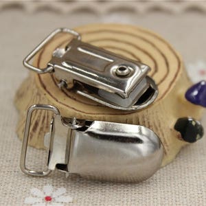 May include: Two silver-colored suspender clips with a rectangular design and a loop. The clips are on a small wooden decorative piece with a light brown color and a few small decorative objects.