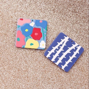 May include: Two square-shaped polymer clay charms. One charm features a floral design with pink, red, yellow, blue, and white flowers on a light blue background. The other charm features a white thorn design on a dark blue background.