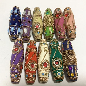 Könnte beinhalten: Zwölf bunte, dekorative Perlen mit goldenen Akzenten und Strasssteinen. Die Perlen sind oval geformt und haben komplizierte Designs.