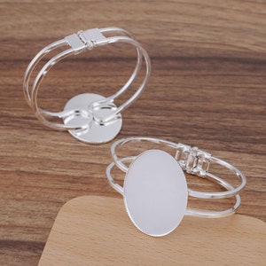 May include: Two silver bangle bracelets with a round, blank, silver charm. The charms are smooth and have a slightly raised edge.