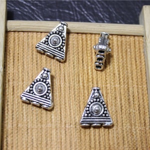 Puede incluir: Cuatro amuletos de metal de forma triangular plateados con diseños ornamentados. Los amuletos se muestran sobre una superficie de madera marrón.