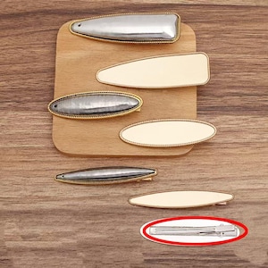 May include: A selection of hair clips in different shapes and colours. The clips have silver and cream-coloured surfaces, some with gold-coloured trim. They are arranged on a wooden surface, highlighting their design. These hair accessories are suitable for various hairstyles and occasions.
