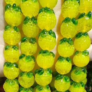 Peut inclure: Gros plan sur une collection de perles de verre jaunes et vertes. Chaque perle est de forme arrondie avec une surface texturée et un accent vert foncé sur le dessus, ressemblant à un petit fruit. Les perles sont disposées en rangées.