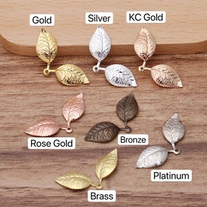 Könnte beinhalten: Sortiment an blattförmigen Metall-Anhängern in verschiedenen Ausführungen, darunter Gold, Silber, Roségold, Bronze, Platin und Messing. Jeder Anhänger besteht aus zwei Blättern, die am Stiel miteinander verbunden sind.