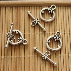 Puede incluir: Cuatro cierres de palanca de metal plateado con diseños ornamentados. Cada cierre presenta un anillo redondo con un patrón decorativo y una barra recta con un pequeño bucle en un extremo.