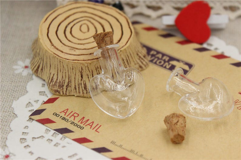 Puede incluir: Dos botellas de vidrio transparente en forma de coraz&oacute;n con tapones de corcho. Las botellas est&aacute;n sobre una superficie de papel marr&oacute;n con el texto "AIR MAIL 00180/2000" impreso en ella.