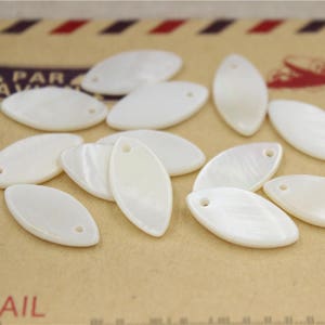 Puede incluir: Una colección de abalorios de concha ovalados de color blanco con un agujero en la parte superior. Los abalorios están dispuestos sobre una superficie marrón.
