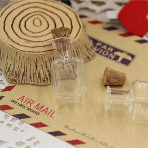 Puede incluir: Dos botellas de vidrio transparente con tapones de corcho. Las botellas est&aacute;n sobre una superficie de papel marr&oacute;n con el texto "AIR MAIL 00190/2000" impreso en ella.