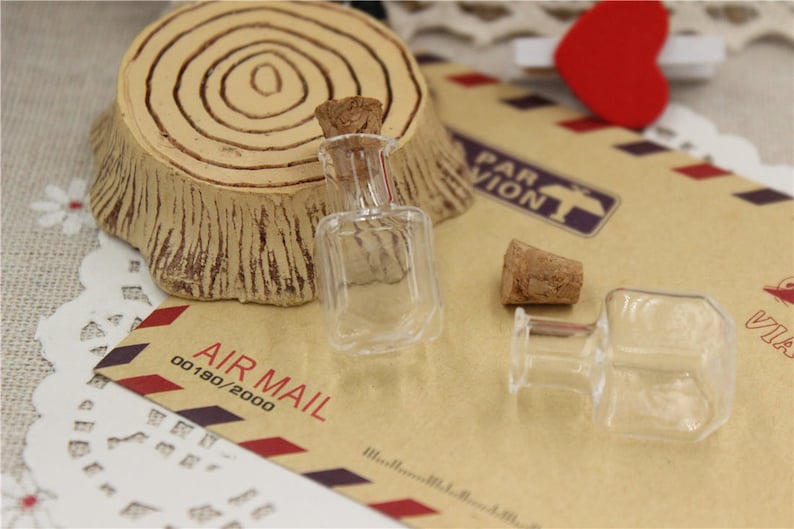 Puede incluir: Dos botellas de vidrio transparente con tapones de corcho. Las botellas est&aacute;n sobre una superficie de papel marr&oacute;n con el texto "AIR MAIL 00190/2000" impreso en ella.