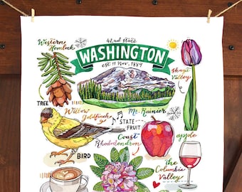 May include: White tea towel featuring a vibrant illustration of Washington State, with images of Mt. Rainier, an apple, a goldfinch, and the Public Market Center. The towel is suspended against a wooden backdrop.