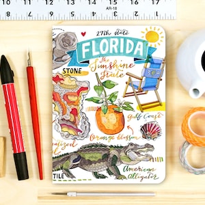 May include: A white notebook with a colorful illustration of Florida. The illustration includes an alligator, an orange blossom, a beach chair, a seashell, and the text "27th state FLORIDA The Sunshine State".