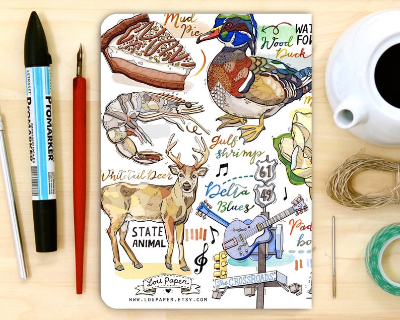 Peut inclure: Un carnet blanc avec une illustration color&eacute;e d'un cerf de Virginie, d'un canard mandarin, d'une crevette, d'une part de tarte &agrave; la boue, d'une guitare et d'une pancarte indiquant "The Crossroads". Le carnet pr&eacute;sente &eacute;galement le texte "State Animal", "Gulf Shrimps", "Delta Blues", "Wood Duck" et "Mud Pie".