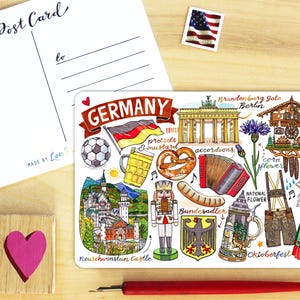 May include: A rectangular postcard featuring a colorful illustration of German cultural icons including the Brandenburg Gate, Neuschwanstein Castle, a nutcracker, a cuckoo clock, a pretzel, a beer stein, and traditional clothing with the text GERMANY.