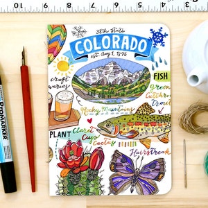 Puede incluir: Una ilustración dibujada a mano de un cuaderno con un fondo de acuarela que representa una cordillera, un pez, una mariposa, un cactus y un vaso de cerveza. La portada del cuaderno presenta el texto "38th State COLORADO est. Aug 1, 1876" y otro texto que incluye "craft weries", "Rocky Mountains", "PLANT", "Claret Cup", "Cactus", "FISH", "green Cutthro Trout" y "Hairstreak".