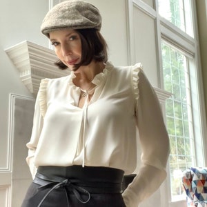 May include: A woman wearing a white blouse with ruffled trim, a brown tweed newsboy cap, a black leather belt, and black trousers.