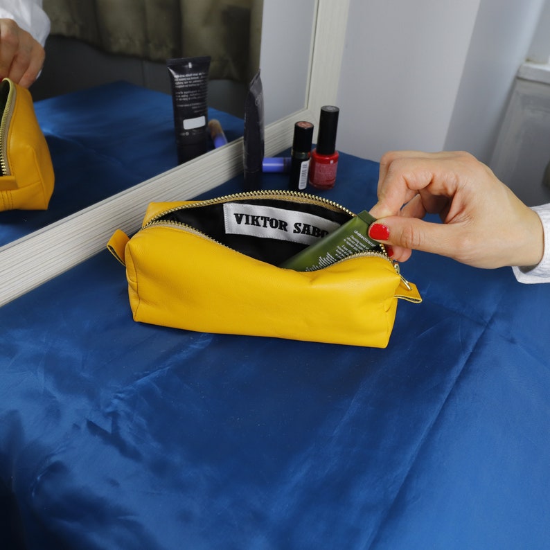 May include: A vibrant yellow leather cosmetic bag with a black interior, open to show a tube of product and a label reading "VIKTOR SABO". The bag sits on a blue surface, with other beauty products in the background.