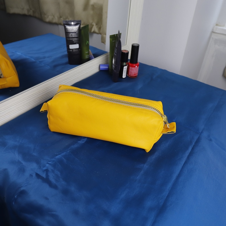 May include: A vibrant yellow rectangular cosmetic bag with a gold zip. The bag is made of a smooth material and rests on a blue surface. Other beauty products are reflected in a mirror behind the bag.