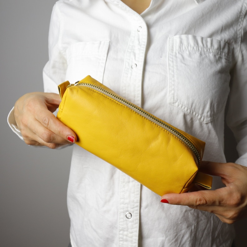 May include: A vibrant yellow rectangular pencil case with a silver zip. The case is made of a textured material and is held by a person wearing a white button-down shirt. The case is approximately 20 cm long.